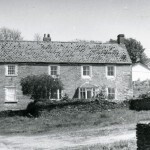 West Wembury farmhouse and yard, 1980. The house is Grade II listed, and is a very substantial stone-built house of the 18th century, with a slate-hung front elevation, granite quoins, and 19th-century sash windows.  This was a working farm for much of the twentieth century, but in the 1960s and 70s its land was almost all developed as today&rsquo;s Wembury was built. At this point the farmstead fell into disuse &ndash; as can be seen from the weeds in the yard and the absence of farm implements and livestock. An equivalent picture of the working farm can be seen in this website&rsquo;s 'Langdon Estate in 1927' photo gallery.  The view is through the farmyard gateway, through which the cows were led twice daily to and from their pasture.  The gateway is now blocked off, and the growth of trees and shrubs has almost obscured the yard and farmhouse from view.